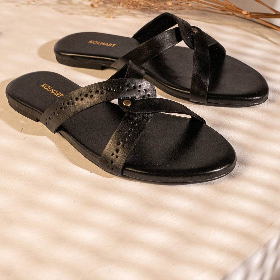 A pair of black leather slide sandals with perforated straps and a small gold-tone button, displayed on a beige surface with dried floral stems in the background.