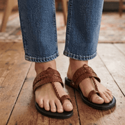 A person wearing brown kolhapuri sandals with black soles, paired with blue jeans, standing on a rustic wooden floor.