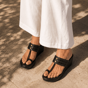 Black woven thong sandals with braided straps and gold-tone studs, worn with white wide-leg pants on a sunlit stone surface.