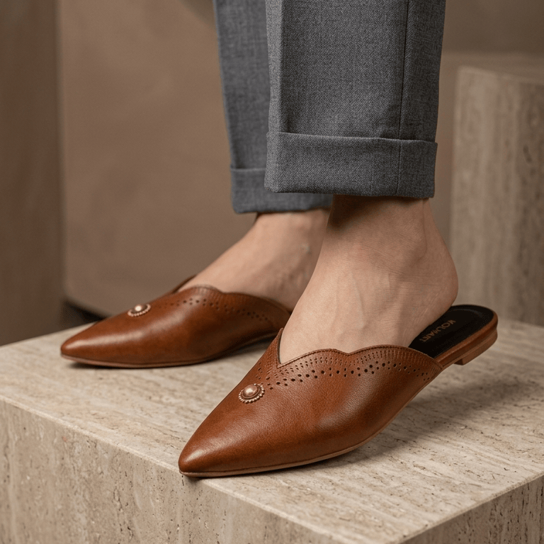 A person wearing brown leather pointed-toe mule flats with gold-tone decorative studs, paired with gray trousers, standing on a beige stone block.