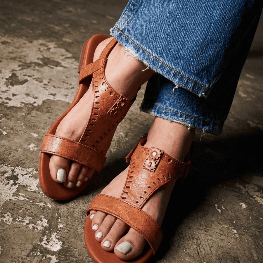 Brown sandals worn with blue jeans on a textured floor.