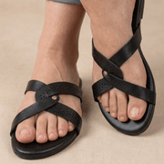 Black leather criss-cross flat sandals worn with cuffed blue jeans, shown on a person's feet against a neutral beige background.