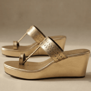 A pair of metallic gold wedge sandals with cutout details on the straps, shown on a neutral beige background.